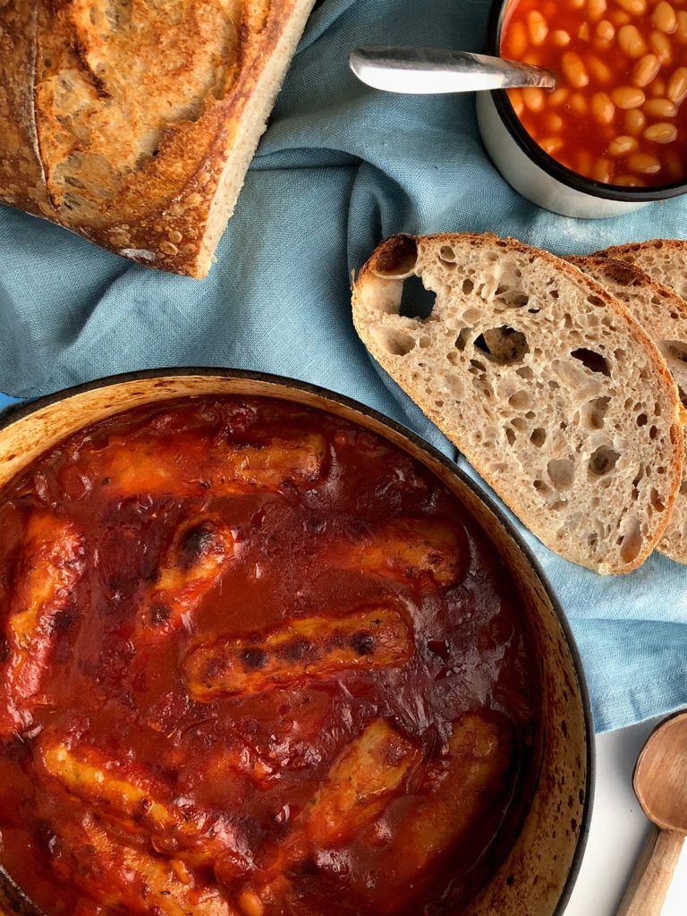 Sausages in red tomato-based sauce in a cast-iron pot, sliced sourdough bread and a mug with baked beans.