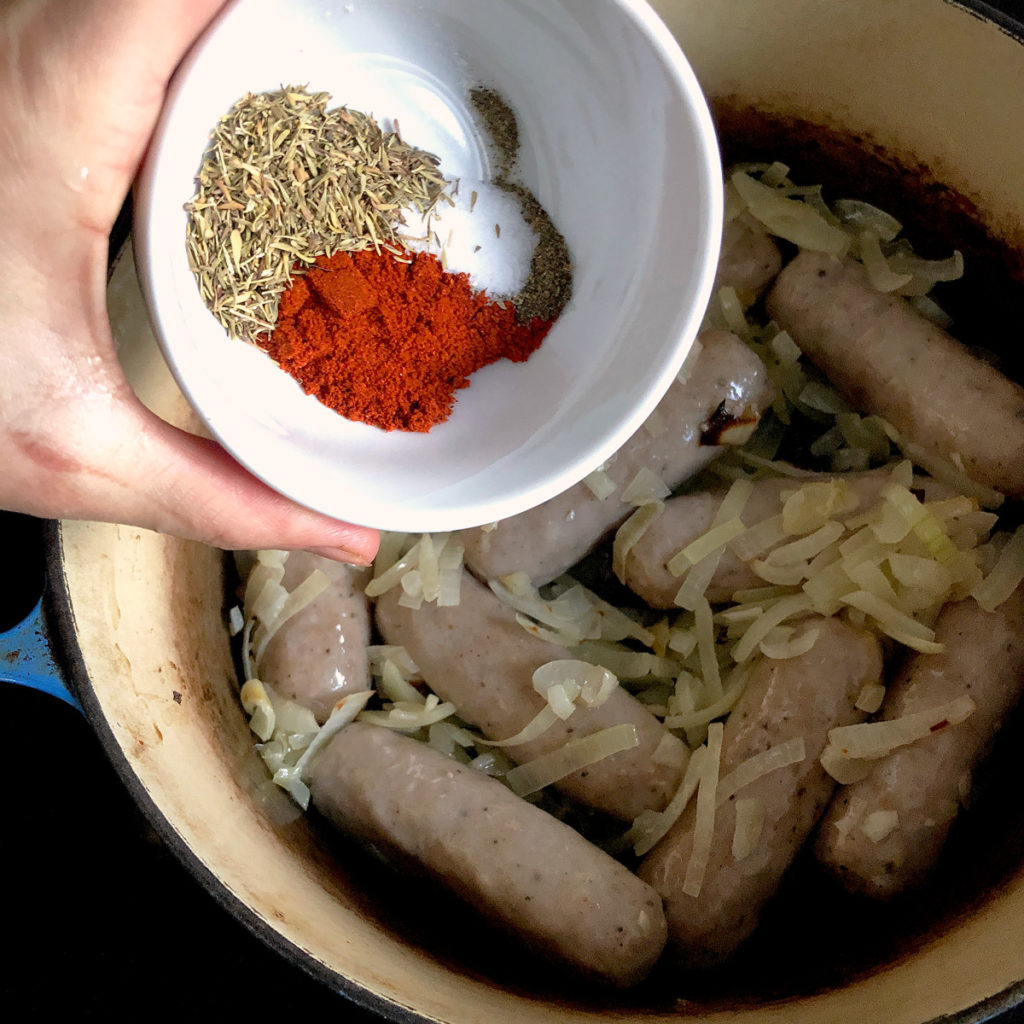 Sprinkling herbs and spices over pre-roasted sausages and onion.