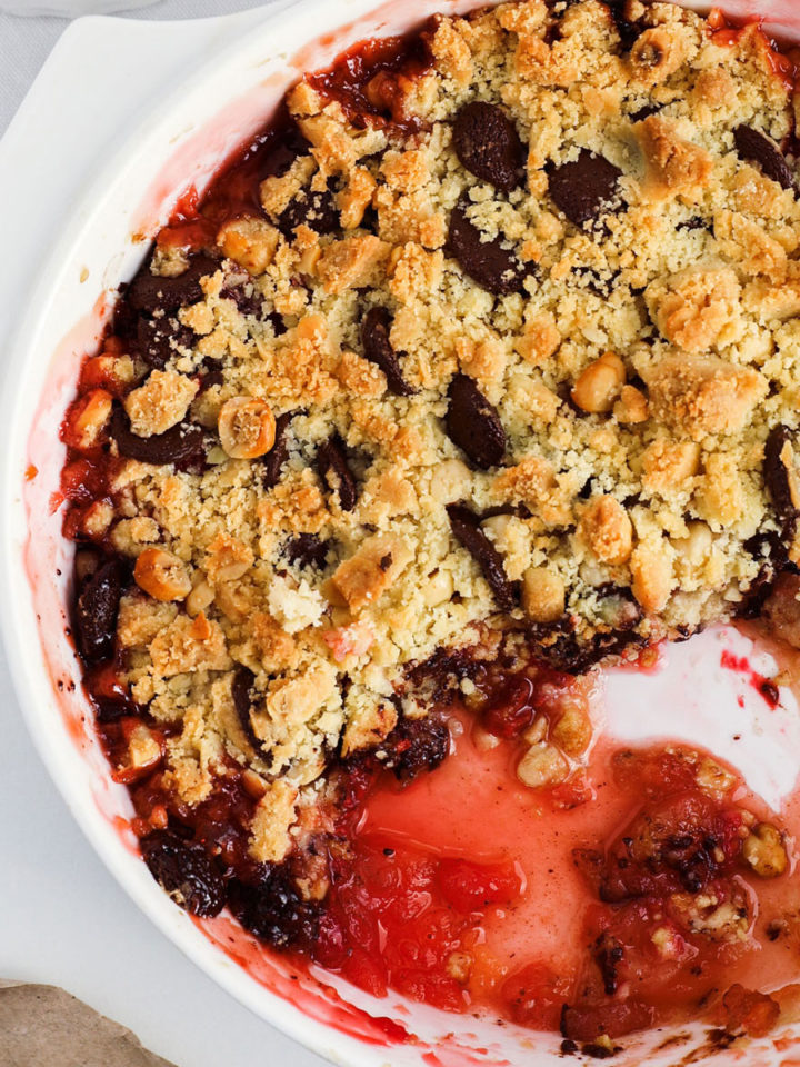 Apple and raspberry crumble with golden lumpy topping and red juices at the bottom in a white round baking dish.