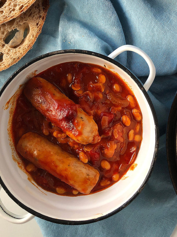 Sausage and bean hotpot in a cast-iron pot, sliced sourdough bread and a portion of hotpot in a white bowl.